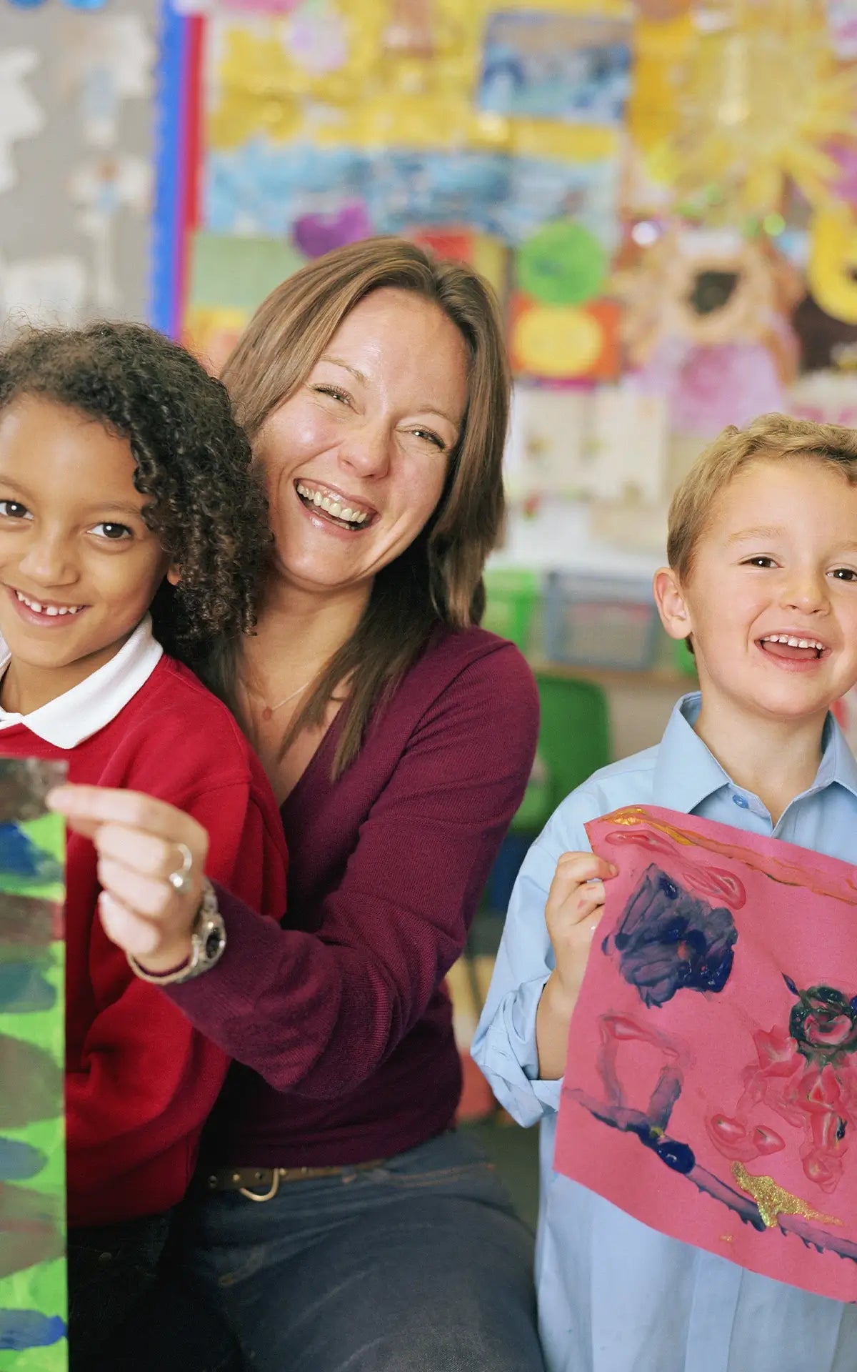 A smiling woman with two children in a colorful art room, holding up children's colorful drawings.